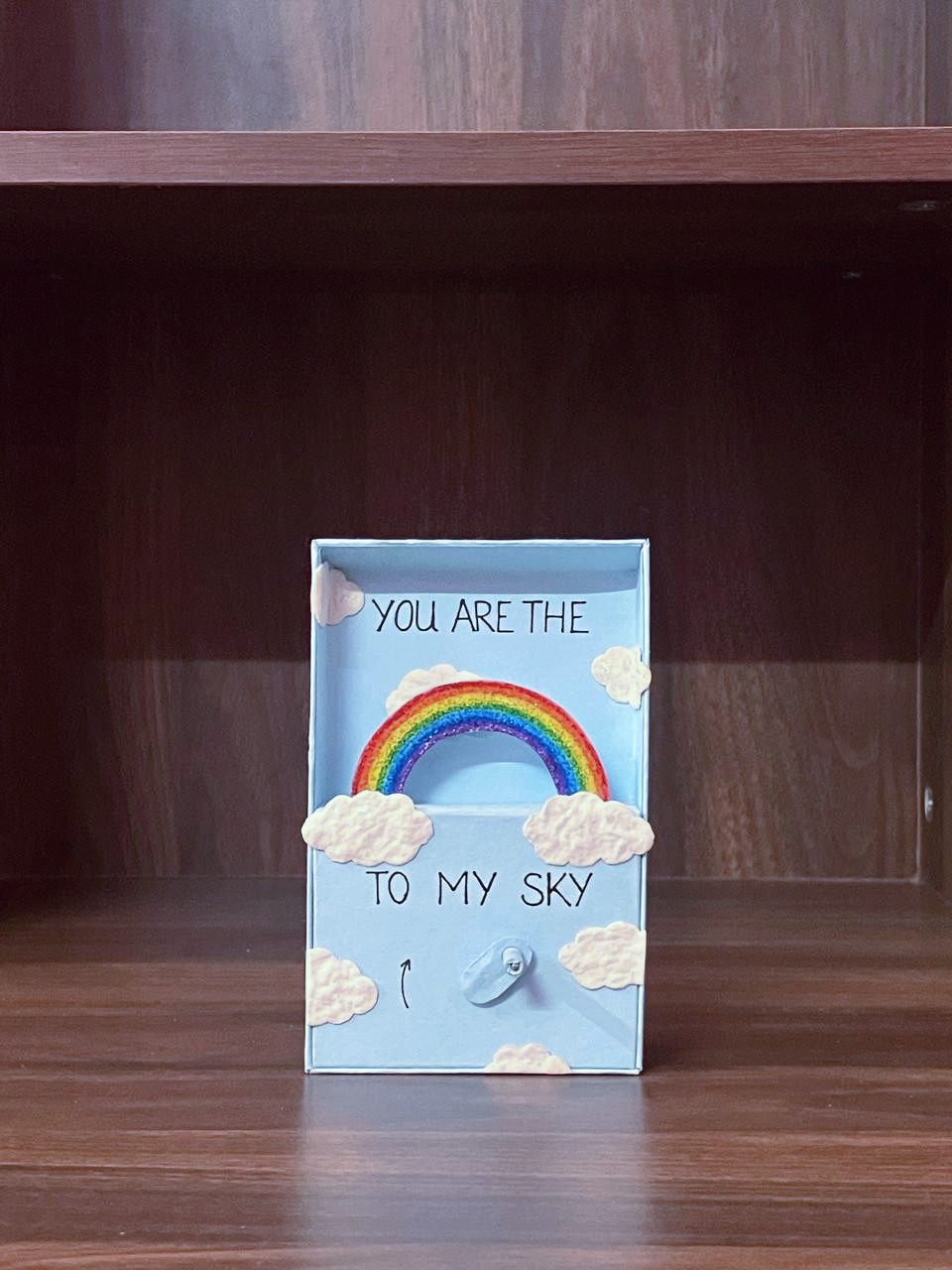 You’re My Rainbow by Shirin Mirza, a small blue automaton with a rainbow rising between clouds, displayed on a wooden shelf.