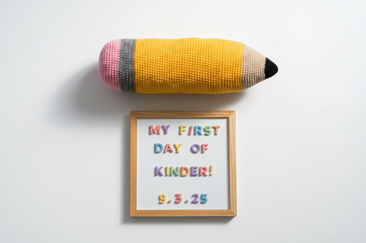 A giant crocheted yellow pencil with a pink eraser lies above a wooden frame reading “MY FIRST DAY OF KINDER! 9.3.25” on a pale surface.