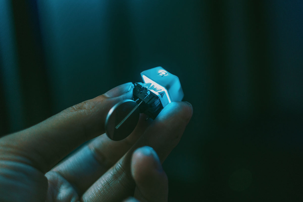 A hand holds a single mechanical keyboard switch and keycap attached to a keyring.