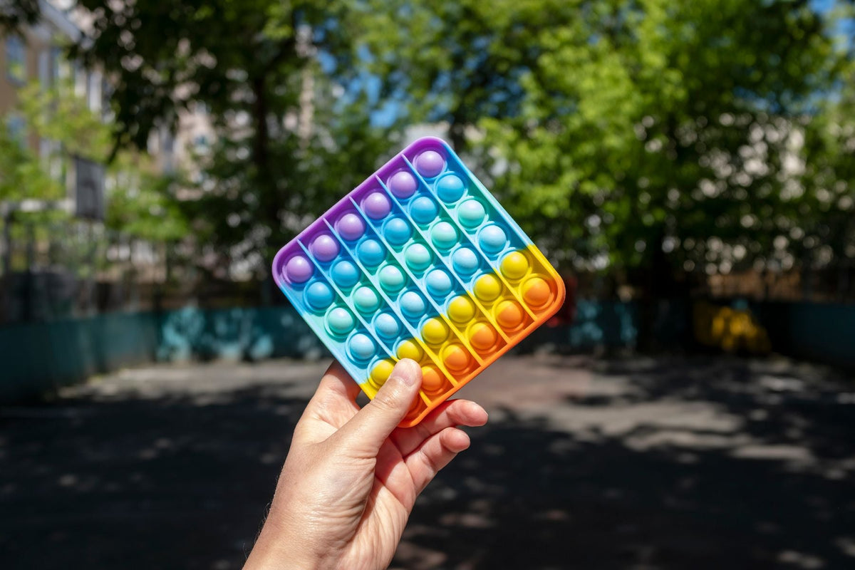 A hand holds a rainbow-colored silicone bubble popper against a blurred outdoor background.