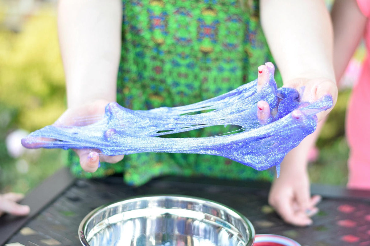 Two hands stretch translucent blue-violet glitter slime into a wide thin sheet outdoors.